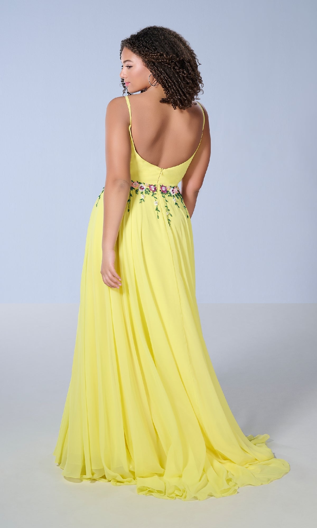 A woman faces away, wearing the Tiffany 16146 Long Plus-Size Prom Dress—a flowy A-line yellow gown with floral beading at the waist—set against a plain light background.